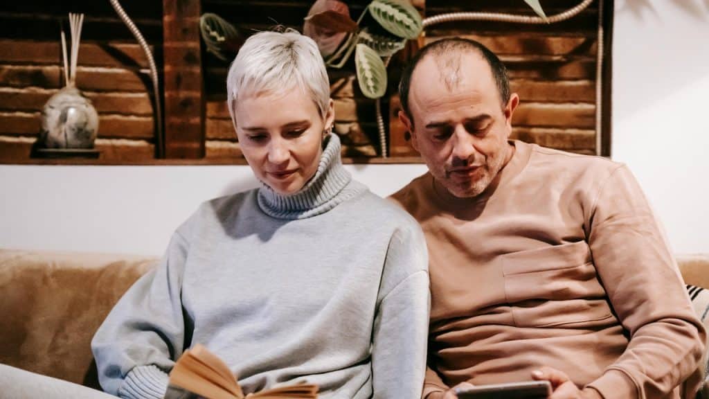 A woman reading a book while a man beside her looks at a tablet.