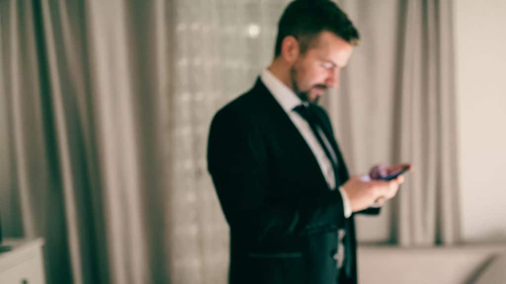 A man wearing a black suit is texting on his cell phone.