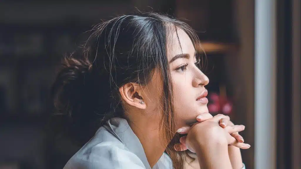 A young woman resting her chin on her hands while gazing thoughtfully out a window.