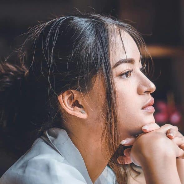 A young woman resting her chin on her hands while gazing thoughtfully out a window.