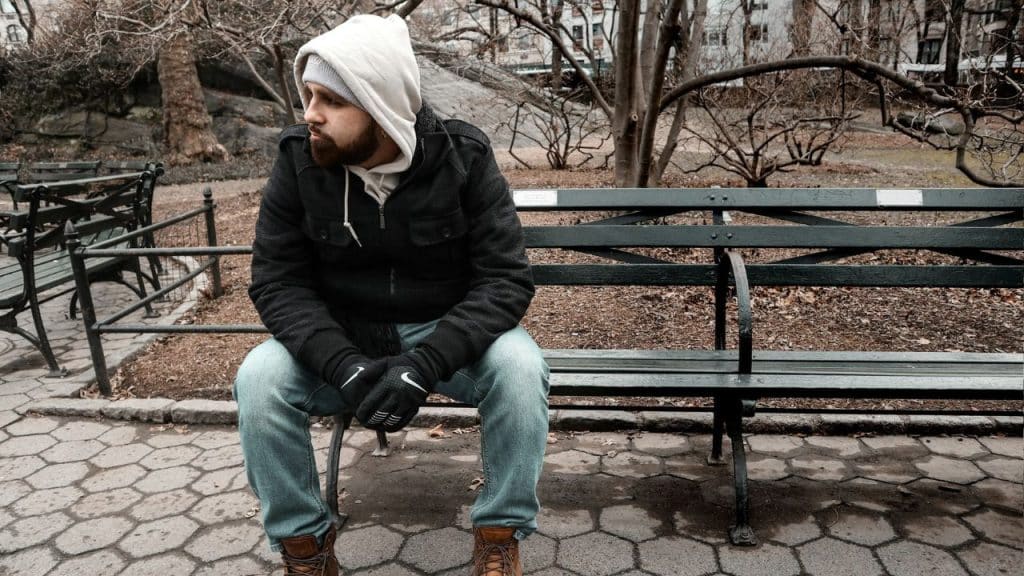 A man in a hooded jacket and gloves sits alone on a park bench.