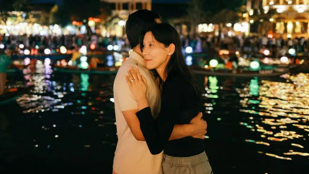 A couple embraces near a water body at night with blurred, colorful lights behind.