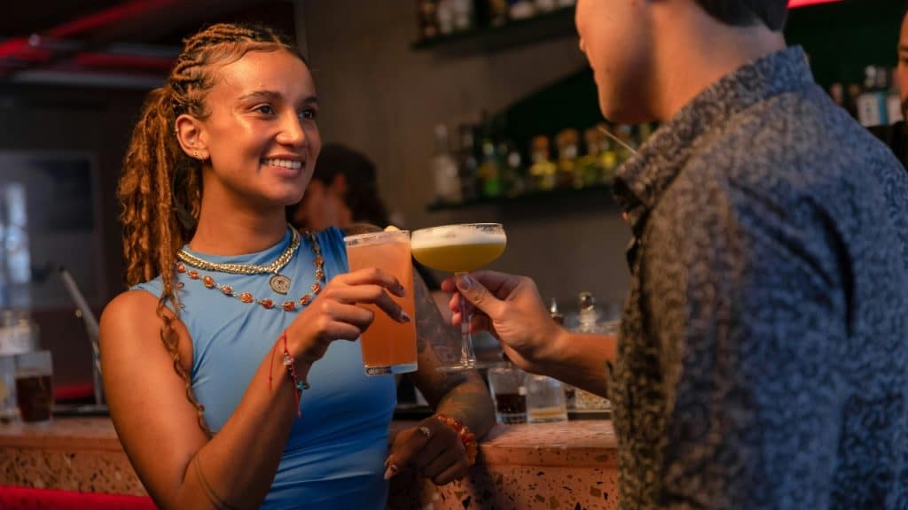 A woman with braided hair smiles and clinks glasses with a man at a bar counter.
