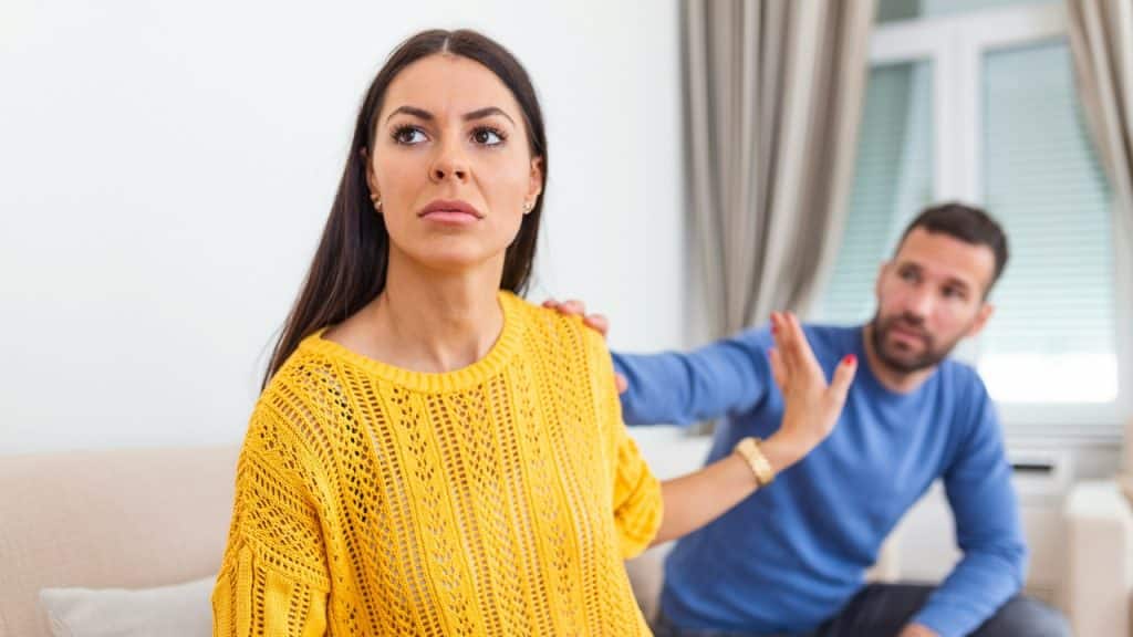 A woman in a yellow sweater gestures as a man reaches toward her shoulder.