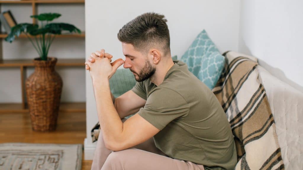 A man sits on a couch with his eyes closed and his hands clasped together.