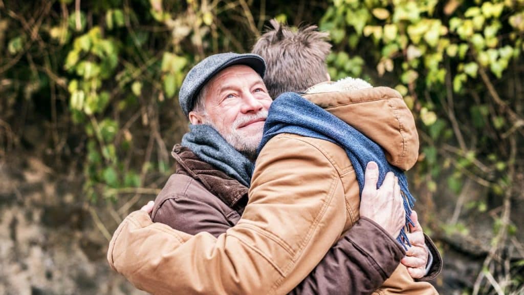 An older man wearing a cap and scarf warmly hugs another person outdoors.