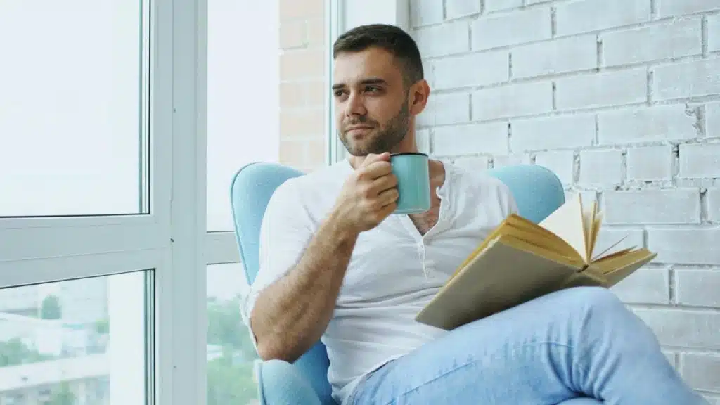 A man in a white shirt sits in a chair, holding a mug and book.