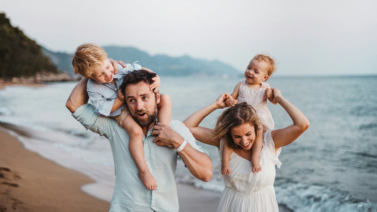 A couple walking down a beach while carrying their kids on their shoulders.