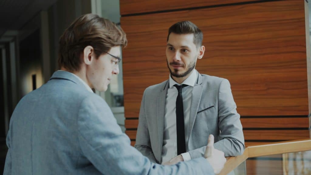 Two men in grey suits stand in a modern hallway while engaged in a conversation.