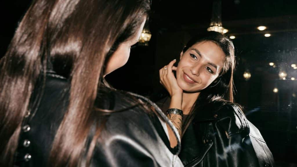 A woman smiling at her reflection in a mirror in a dimly lit room.