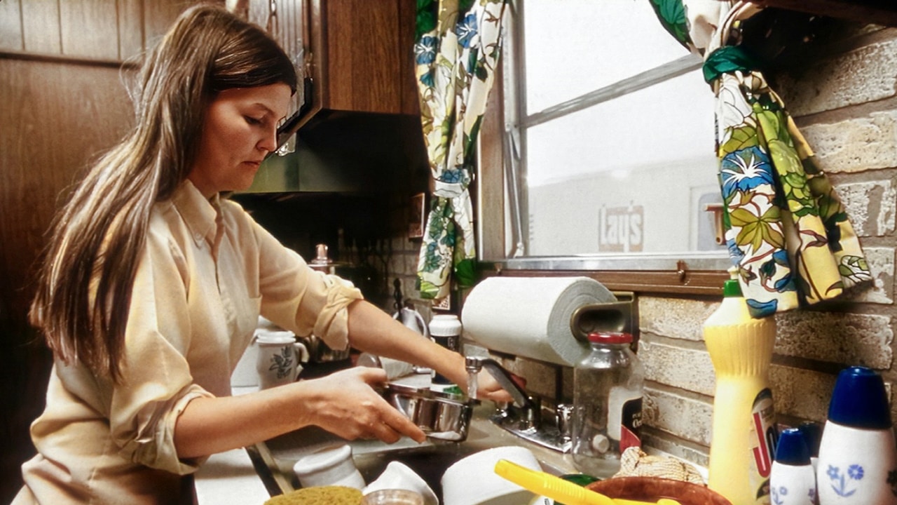 Woman washing dishes at a sink in a retro kitchen with wooden cabinets and a brick wall