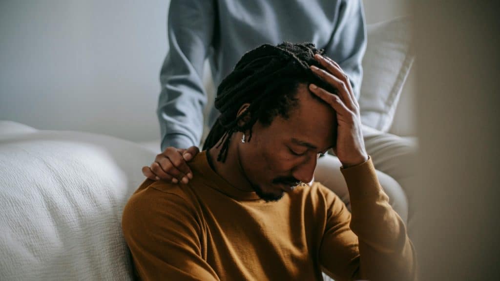 A woman places a comforting hand on the shoulder of a man looking down.