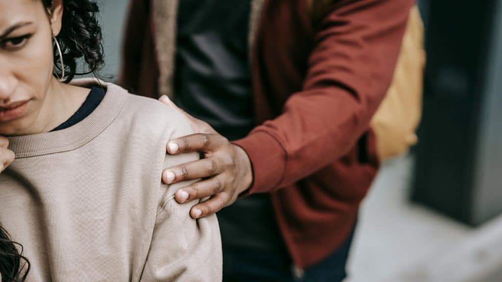A person in a red jacket places a hand on a woman's shoulder.