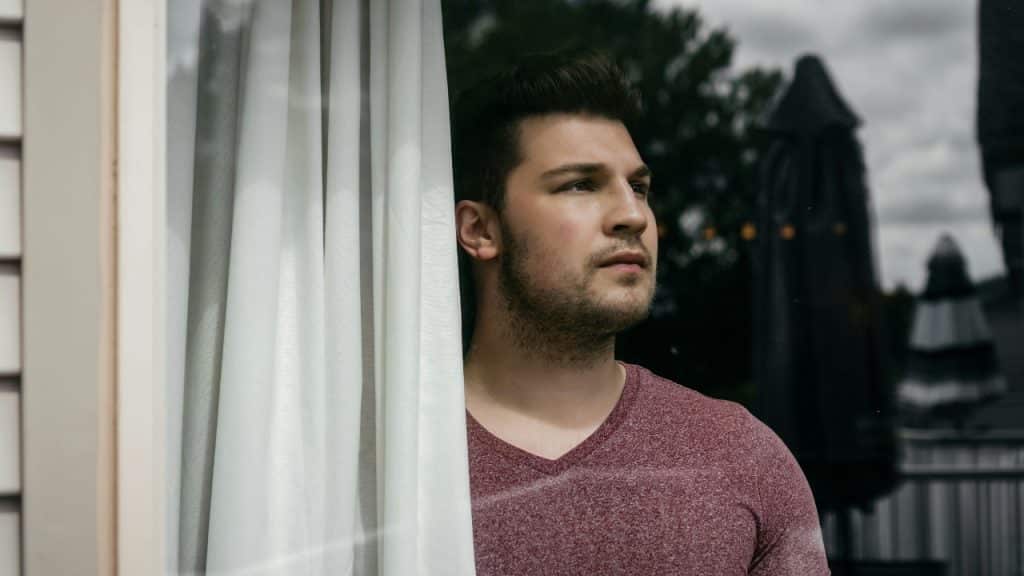 A man in a maroon shirt looks out a window next to a white curtain.