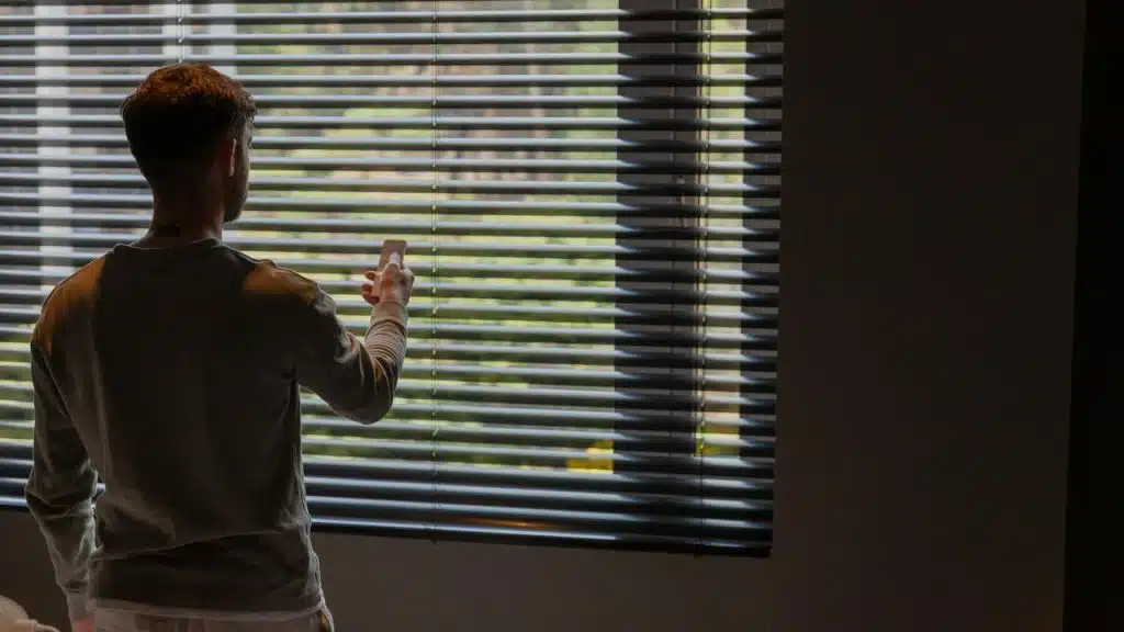 A man using a smartphone while standing by window blinds.