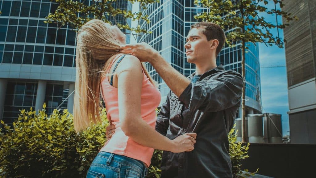 A man reaches toward a woman’s neck while they stand outdoors near a tall building.