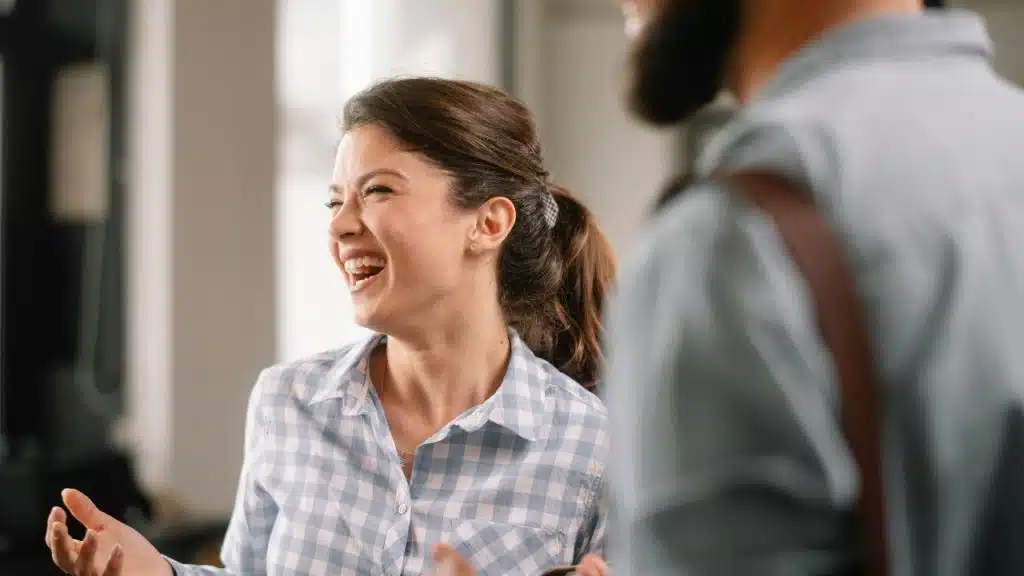 A woman in a checkered shirt laughs with her hands up while facing another person.