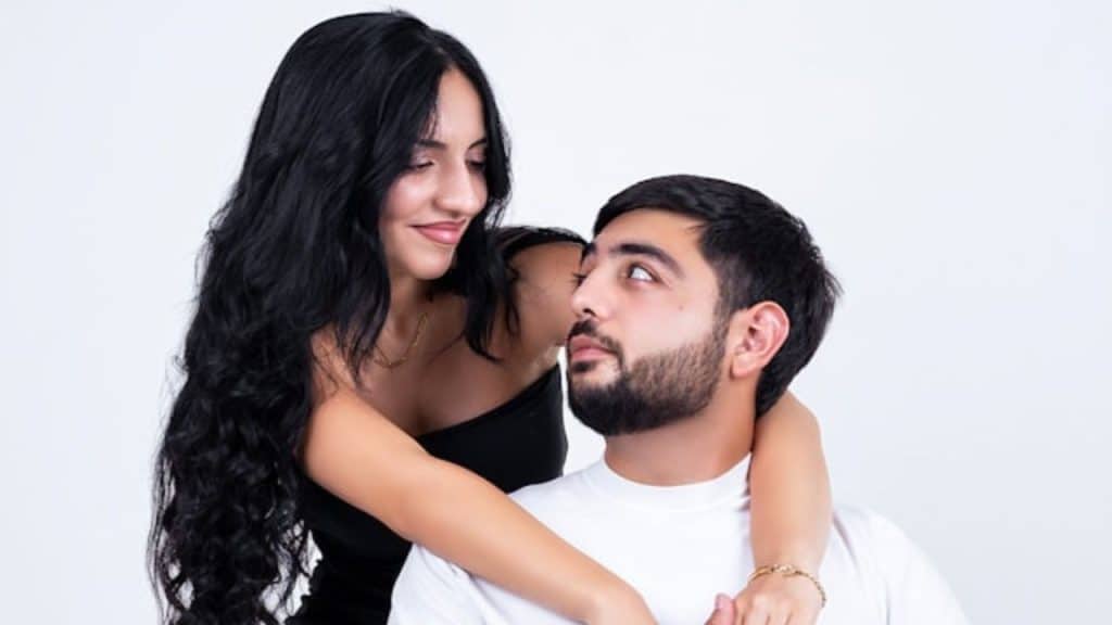A woman with long dark hair leaning over a man sitting in a white shirt.