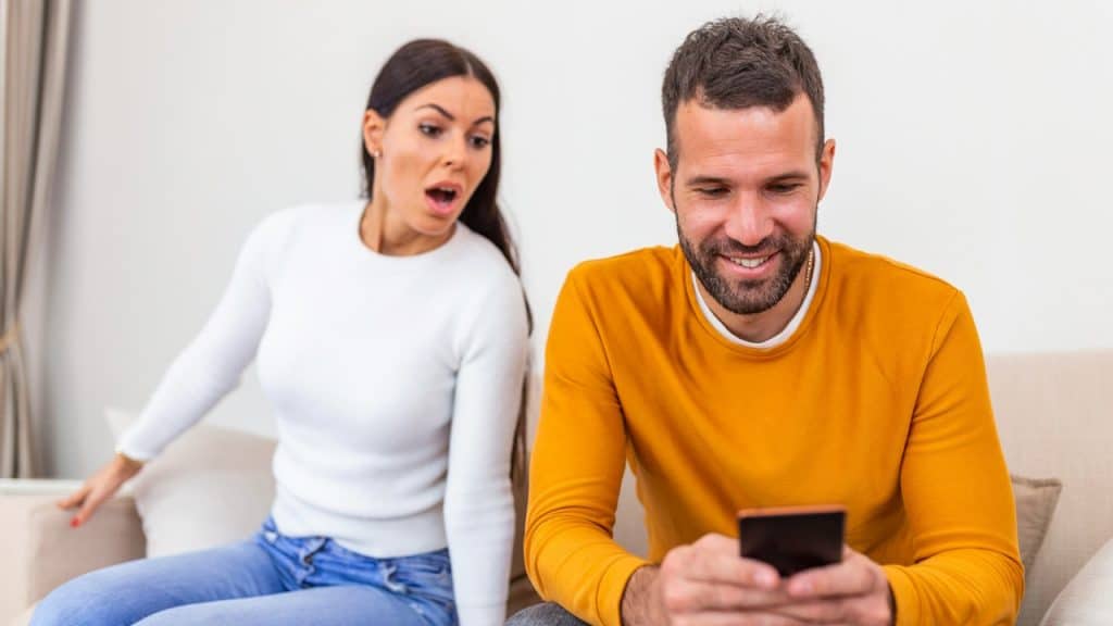 A man is smiling while using his phone, and his wife is looking at the screen from behind in shock.