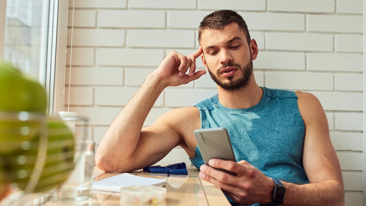 A man in a blue tank top looks at a smartphone while resting his temple on his finger.