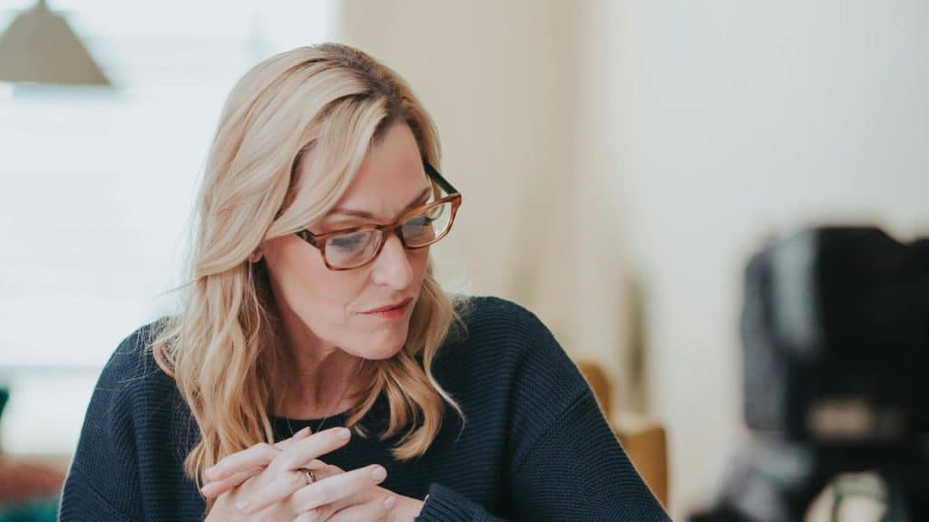 A woman wearing glasses looks down while speaking with hands clasped.