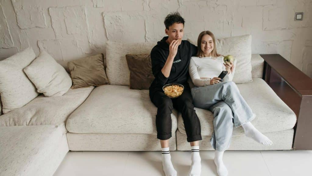 A couple sitting on a couch, watching TV together and eating snacks.