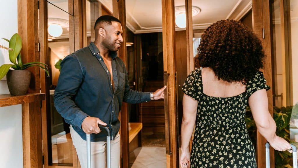 A man holding a suitcase opens a wooden door for a woman also carrying a suitcase.