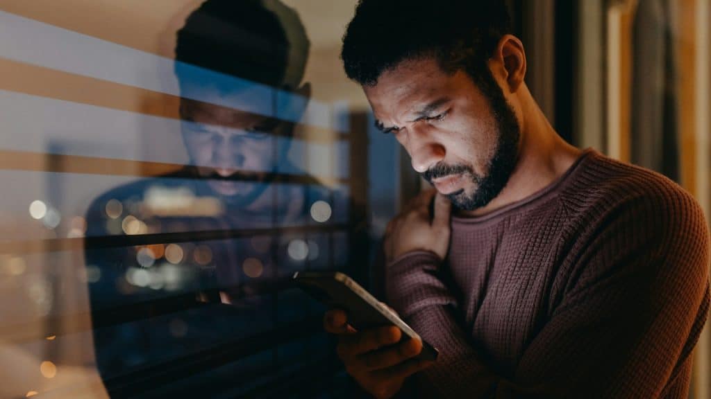 A man looks down at a glowing smartphone at night while his reflection appears in glass.