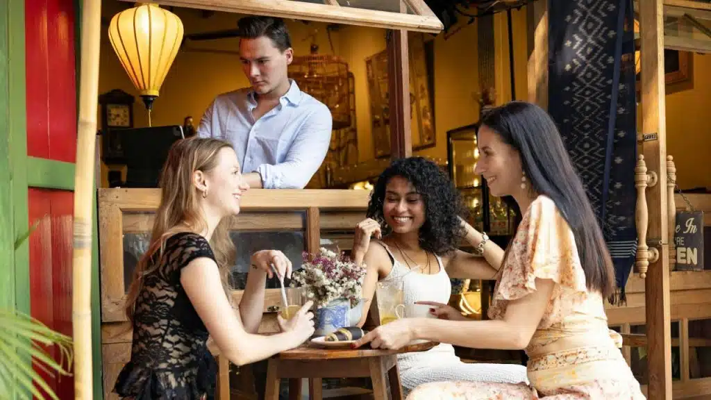 Three women sit smiling at a cafe table while a man stands behind a counter.