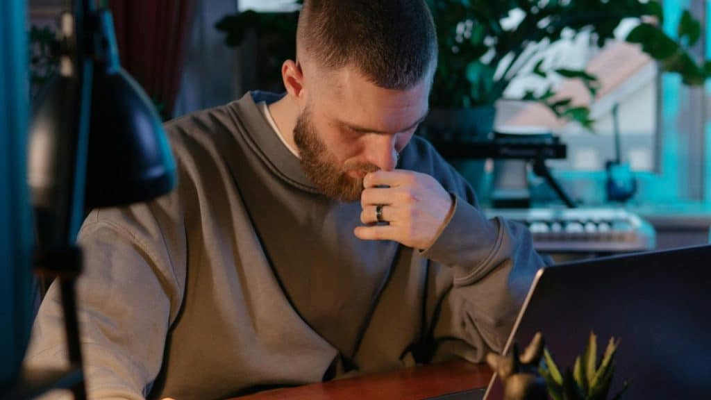 A bearded man sitting at a desk, looking down with his hand near his mouth.