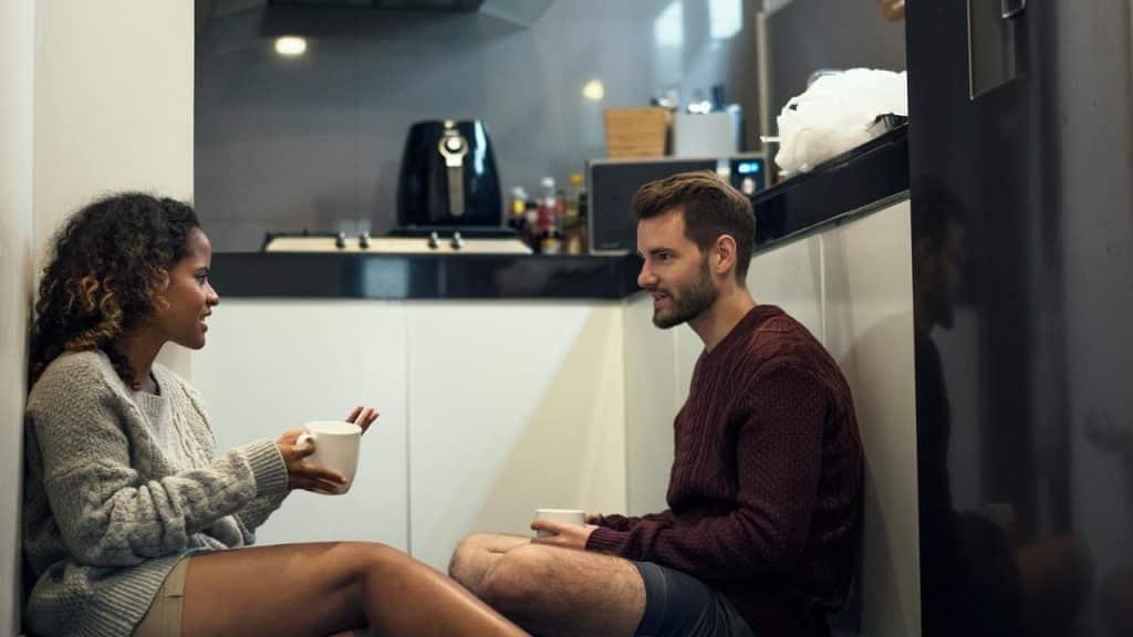 A couple sitting on a kitchen floor, talking quietly while holding mugs.