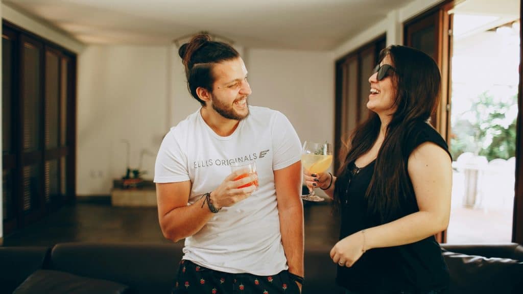 A man and a woman stand together indoors laughing and holding glasses with colorful drinks.