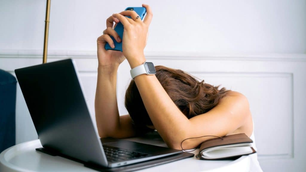 A person rests their head on a desk next to a laptop while holding a smartphone.