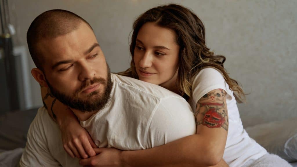 A woman with a tattoo on her arm hugs a man from behind indoors.