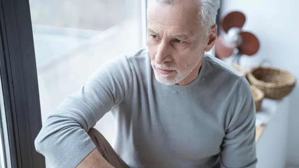 An older man with a white beard looks out a window with a serious expression.