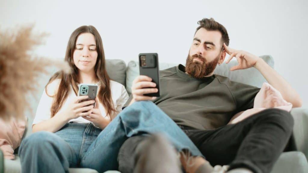 A man and woman sit on a couch, both focused on their individual mobile phones.