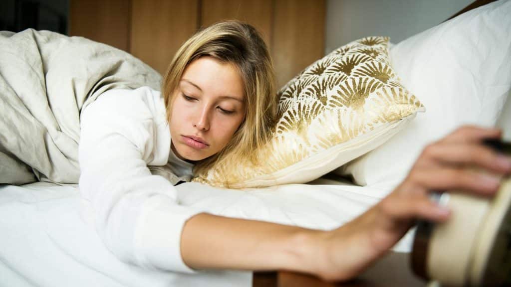 A sleepy woman reaching to turn off an alarm while lying in bed.