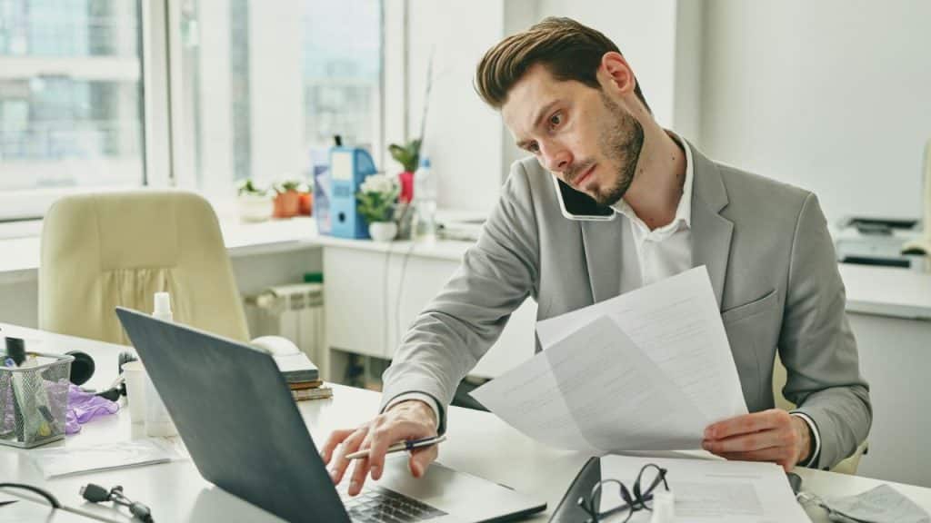 A man in a suit talks on a phone while typing on a laptop at work.