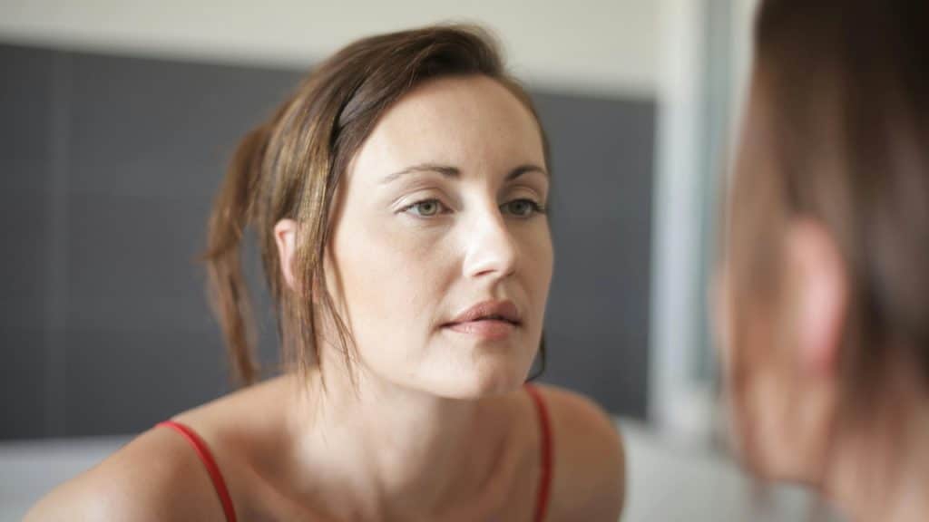 A woman with her hair pulled back looks directly into a mirror at her reflection.