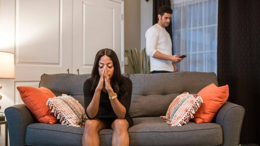 A woman sits on a sofa with hands together while a man uses a phone.