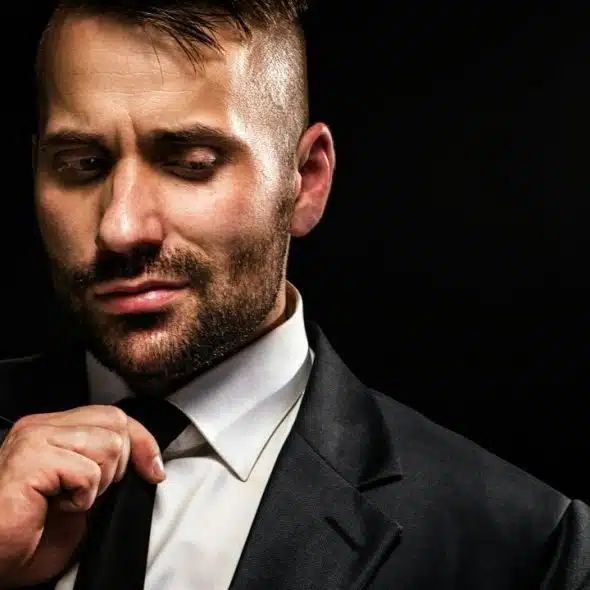 A man with short hair and a beard adjusts his black tie against a dark background.