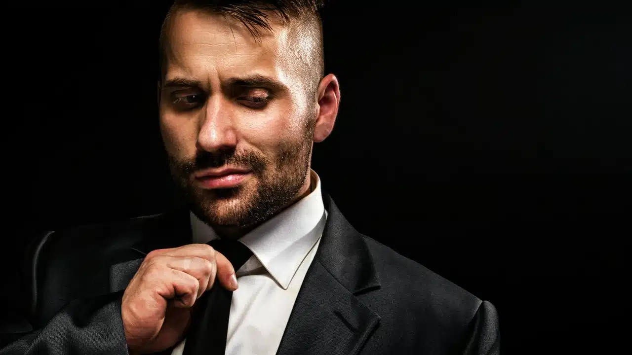 A man with short hair and a beard adjusts his black tie against a dark background.