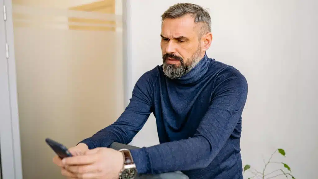 A man with a grey beard looks at a smartphone while wearing a blue turtleneck.
