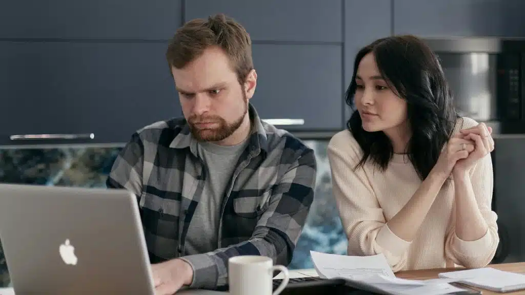 A man and woman look at a laptop screen together with papers on the table.
