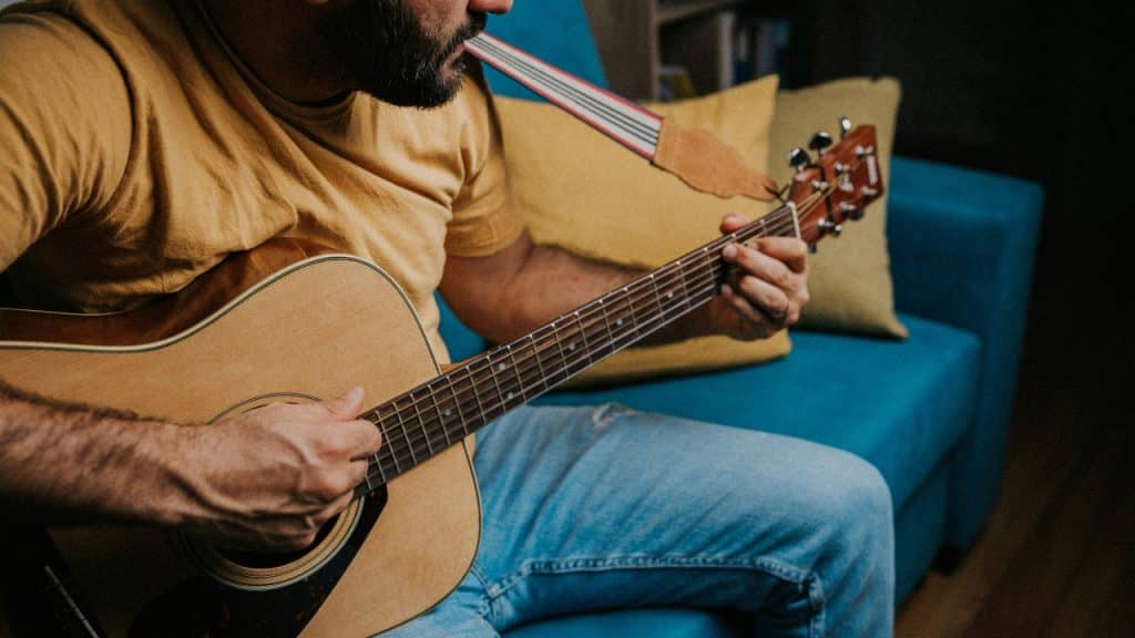 A person in a yellow shirt plays an acoustic guitar while sitting on a sofa.