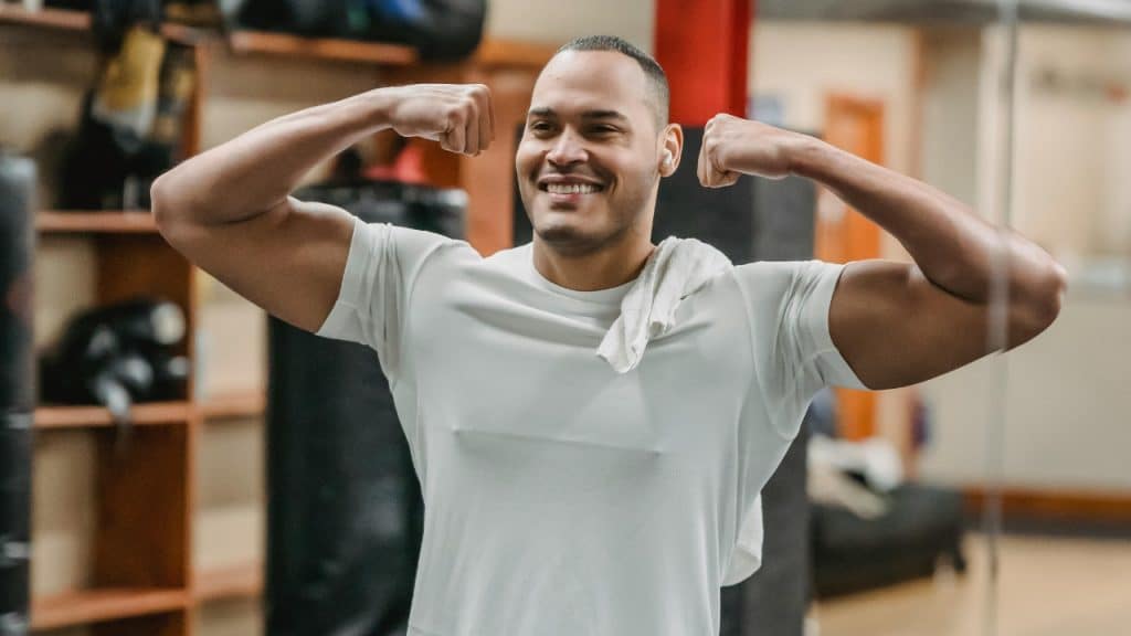 A man in a white t-shirt smiles while flexing his biceps in a gym locker room.