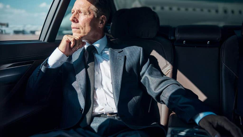 A man in a grey suit and tie sits in the back of a car looking out.