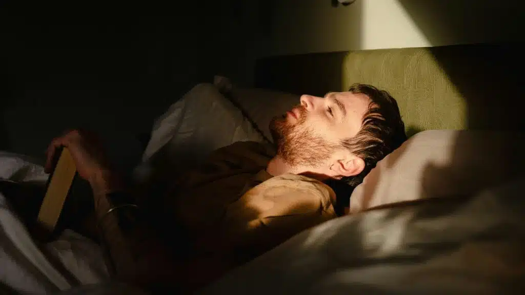 A man with a beard lies in bed with his eyes closed while holding a book.