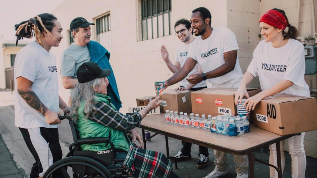 Volunteers give supplies to a man in a wheelchair at an outdoor table.