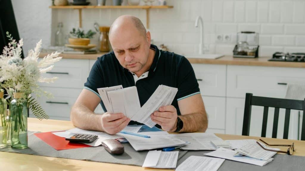 A bald man sits at a kitchen table sorting through many paper documents.